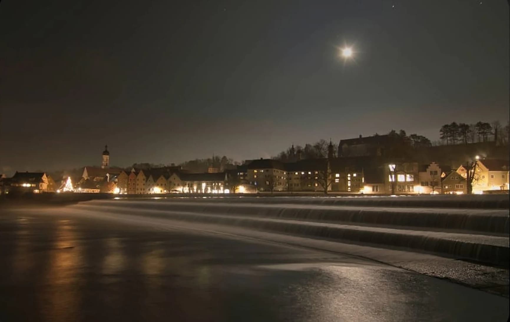 Landsberg am Lech bei Nacht: das Lechwehr im Mondlicht, dahinter die beleuchtete Altstadt