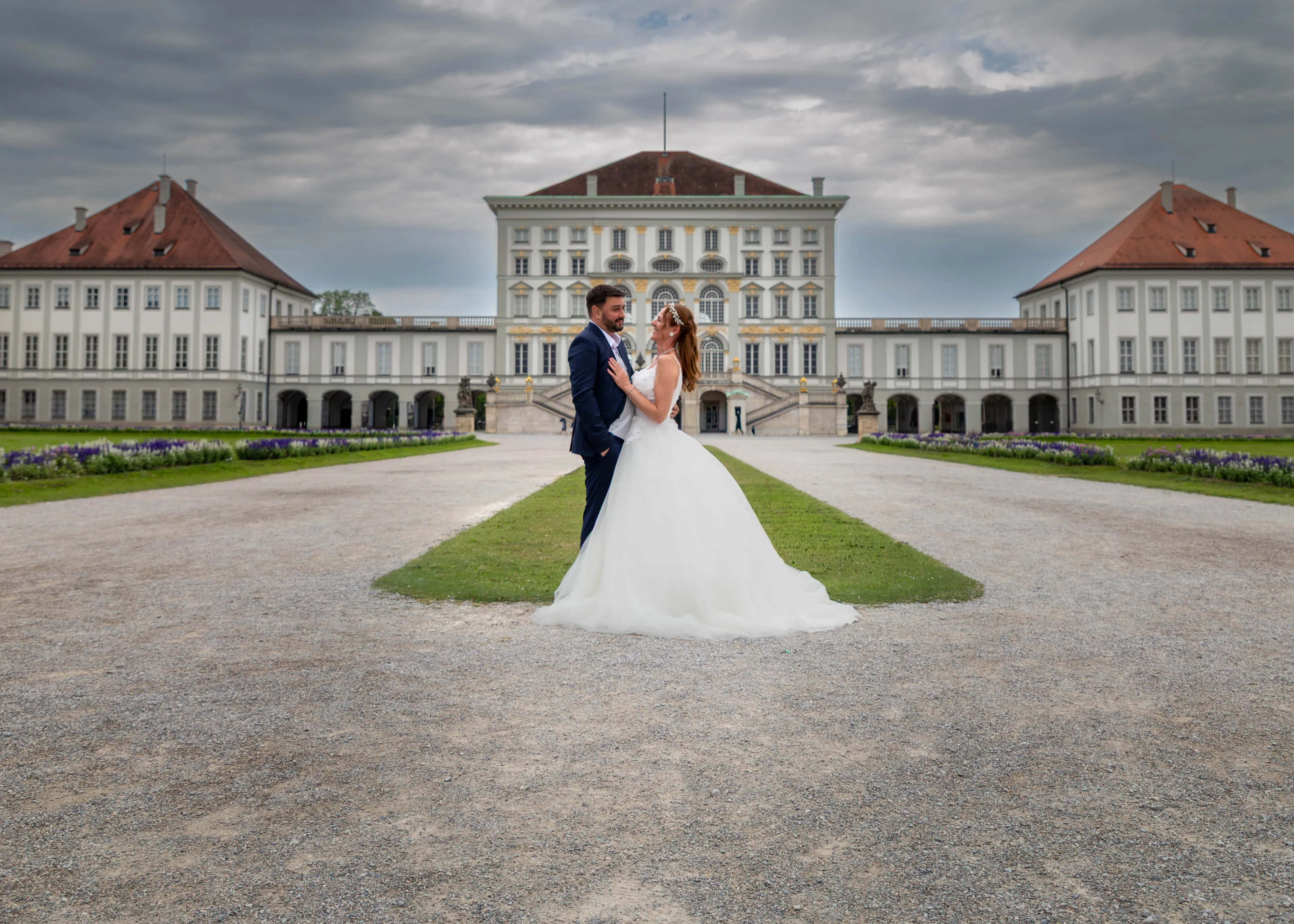 Brautpaar vor dem Schloss Nymphenburg in München, hinter ihnen die weiten Flügel der Schlossanlage unter Wolkenhimmel